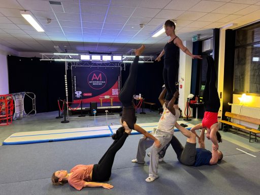 Sechs Personen führen eine Akrobatikübung auf einer Matte in einem Sportraum durch.